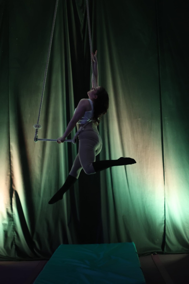Andrea in an arabesque above the trapeze bar in front of a green curtain